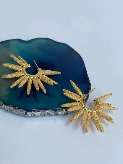 A pair of gold-colored textured sun ray Hoop  earrings displayed on a blue agate stone.