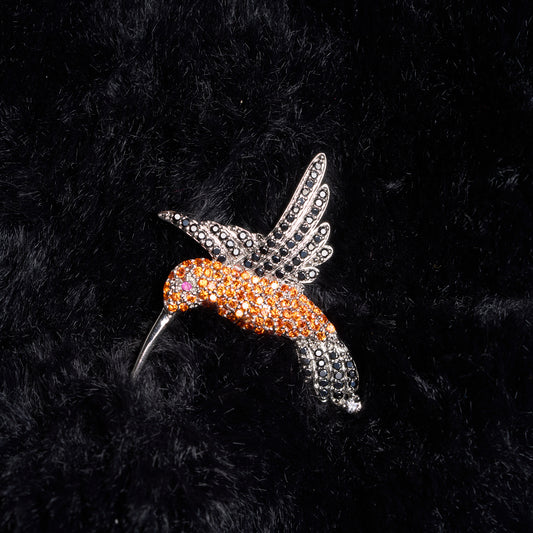 Decorative hummingbird brooch with orange and silver design on a black textured background