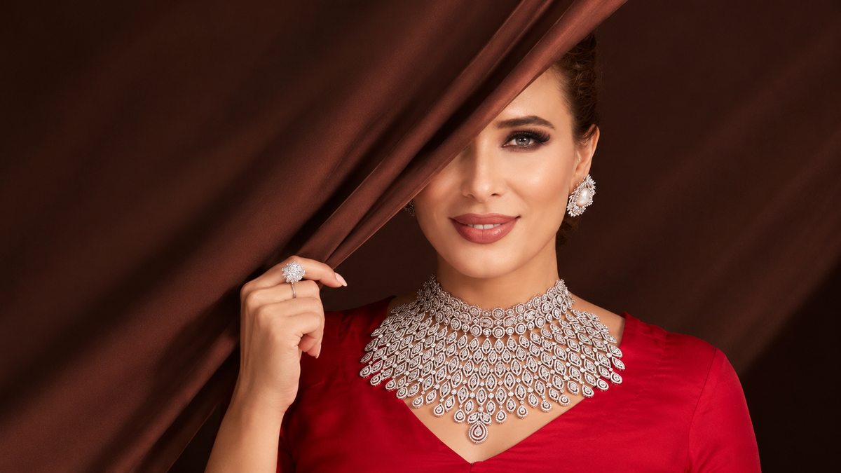 A woman in a red dress wearing an elaborate diamond necklace and matching earrings, holding back a dark brown curtain with one hand.